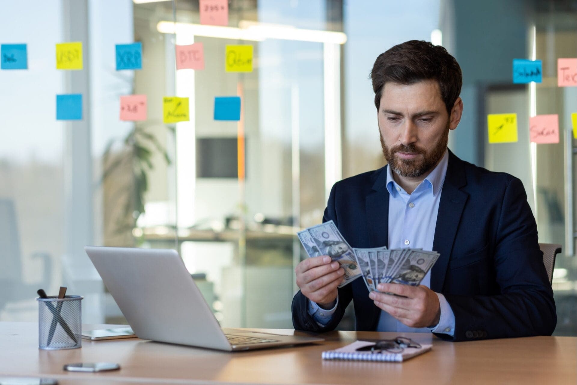 Businessman counting money at desk in modern office with laptop and notes on glass wall, reflecting professional finance management